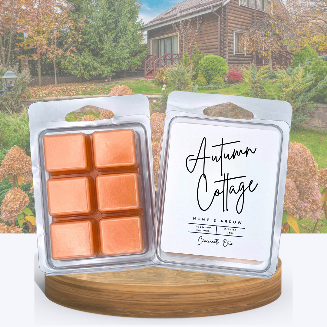 Autumn Cottage soy wax melts in clamshell packaging, displayed on a wooden stand with a cozy log cabin and fall garden in the background.