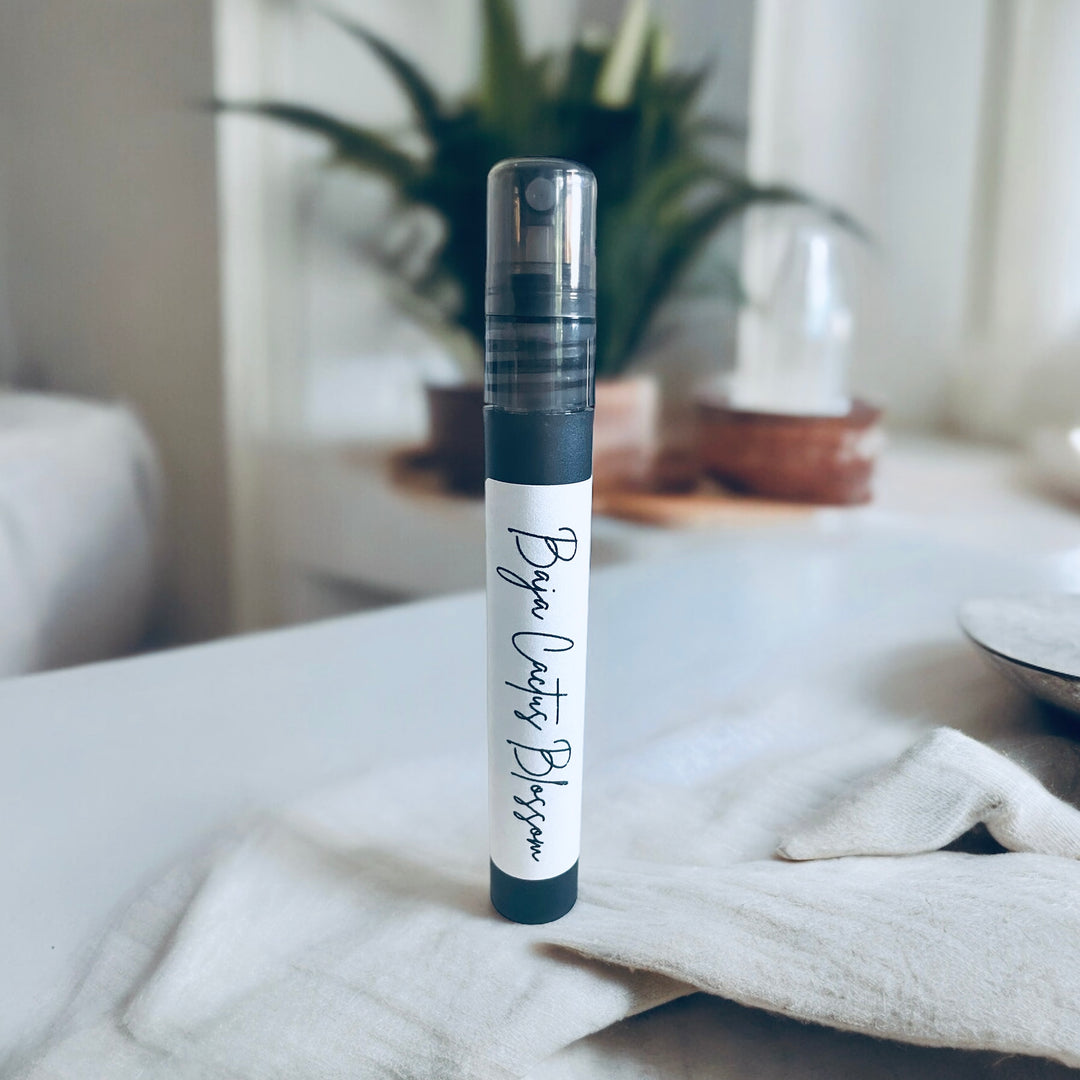 A 10ml room spray bottle labeled 'Baja Cactus Blossom' placed on a white surface with a plant in the background.