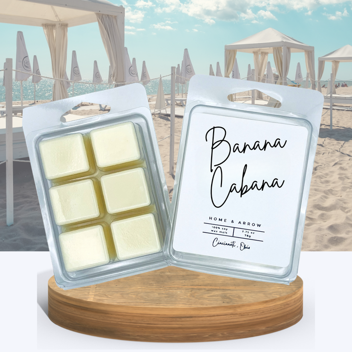 Banana Cabana creamy wax melts in clamshell packaging, displayed on a wooden stand with sand and beach cabanas in the backgrougnd.