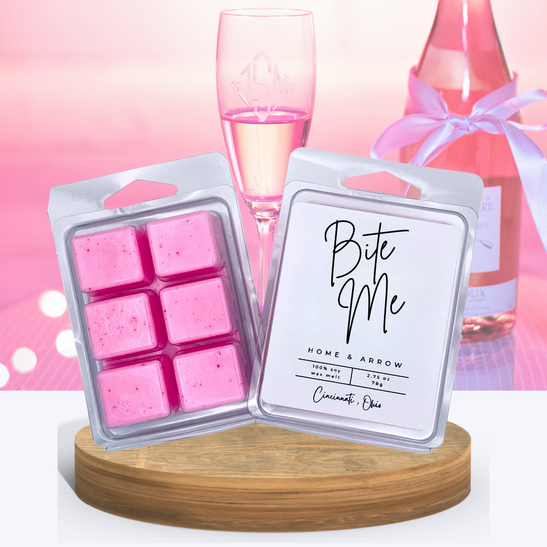 Bite Me pink wax melts in clamshell packaging, displayed on a wooden stand with pink Champagne wrapped in ribbon in the background.