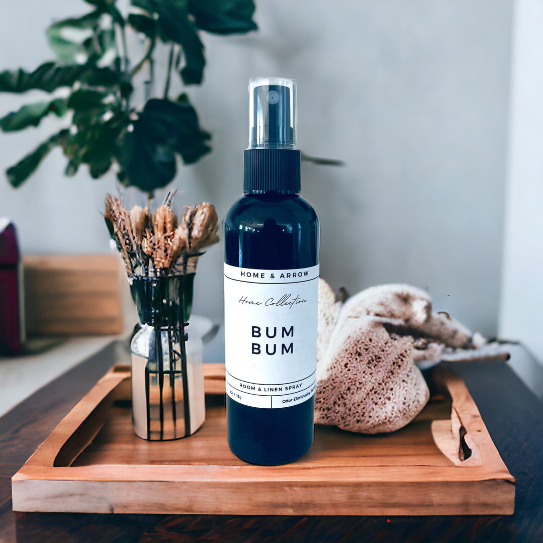 A black spray bottle labeled 'BUM BUM' placed on a wooden tray with a plant in the background and a decorative vase with dried plants to the side.