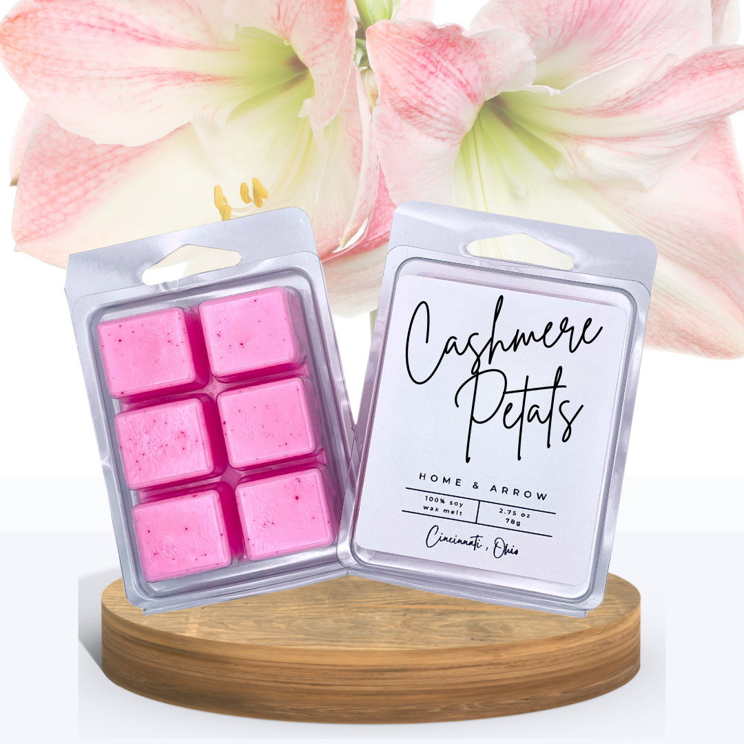 Cashmere Petals wax melts in clamshell packaging, displayed on a wooden stand with beautiful pink and white flowers in the background.