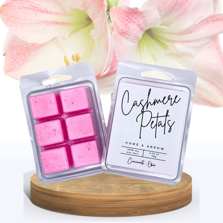 Cashmere Petals wax melts in clamshell packaging, displayed on a wooden stand with beautiful pink and white flowers in the background.