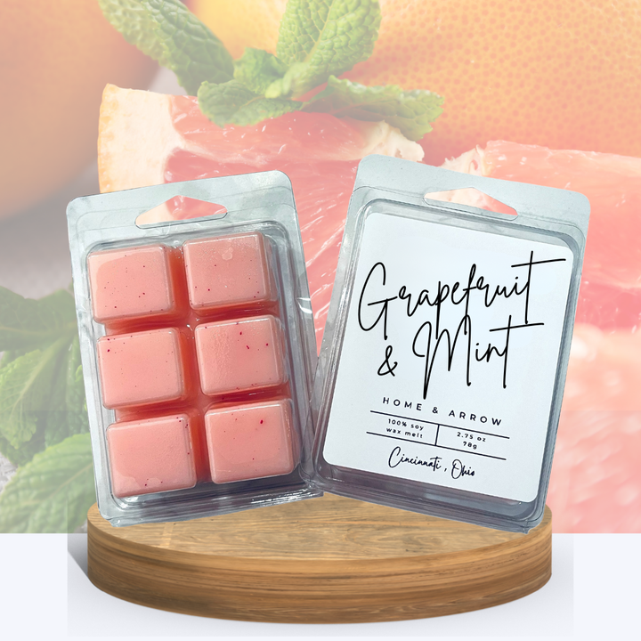 Grapefruit & Mint pink speckled wax melts in clamshell packaging, displayed on a wooden stand with a wedge of grapefruit topped with mint in the background.