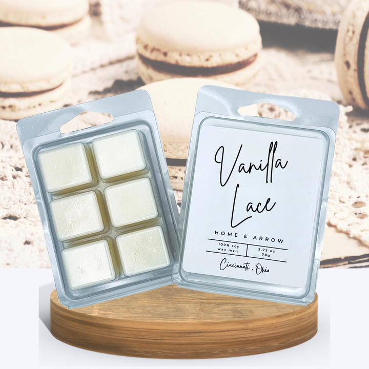 Vanilla Lace creamy wax melts in clamshell packaging, displayed on a wooden stand with several vanilla macaron cookies on lace in the background.