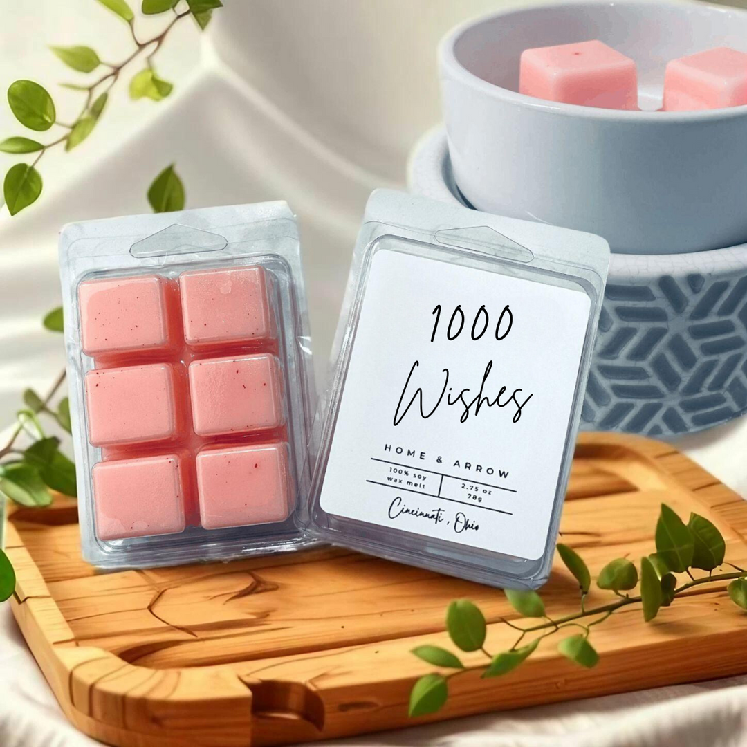 Pink wax melt clamshell labeled '1000 Wishes' sitting on a wooden surface with a wax warmer and greenery in the background.