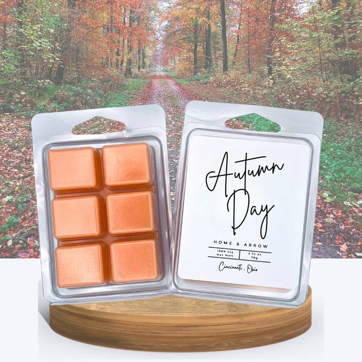 Autumn Day soy wax melts in clamshell packaging, displayed on a wooden stand with a colorful fall forest path in the background.