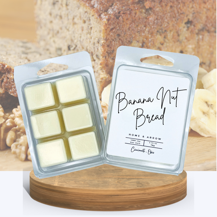 Banana Nut Bread soy wax melts in a clamshell package dispayed on a wooden stand with a cut loaf of banana nut bread in the background with bananas and walnuts at the side. 