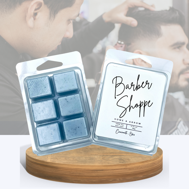 Barber Shoppe soy wax melts in clamshell packaging, displayed on a wooden stand with a man in a getting his hair cut in the background.