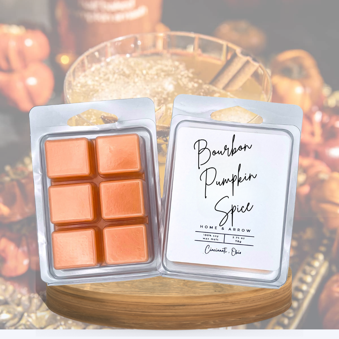 Two wax melts in clear plastic clamshell packaging displayed side by side on a wooden board. The left package contains six orange-brown wax cubes, and the right package has a white label reading 'Bourbon Pumpkin Spice' with 'Home Arrow' and 'Cinnamon Rub' printed on it.
