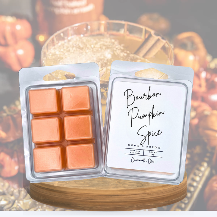 Two wax melts in clear plastic clamshell packaging displayed side by side on a wooden board. The left package contains six orange-brown wax cubes, and the right package has a white label reading 'Bourbon Pumpkin Spice' with 'Home Arrow' and 'Cinnamon Rub' printed on it.