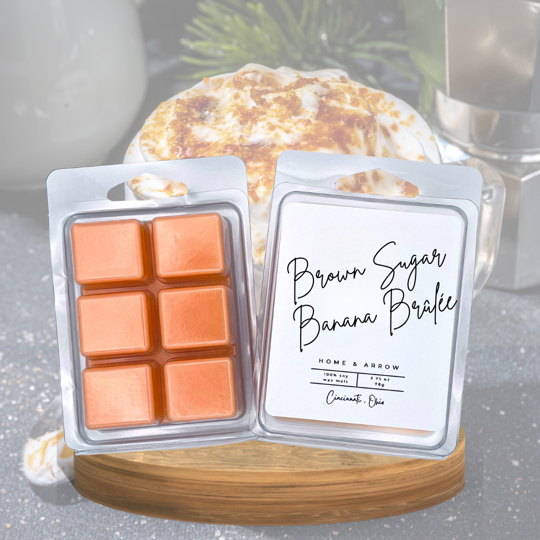Two wax melts in clear clamshell packaging displayed on a wooden board. One side shows six orange-brown wax cubes, and the other side shows a white label reading 'Brown Sugar Banana Butter' with 'Home & Arrow' branding.