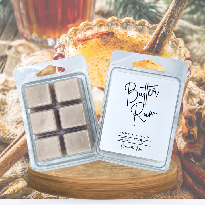 Two wax melts in clear plastic clamshell packaging, one showing six beige wax cubes and the other showing the front label that reads 'Butter Rum' and 'Home & Arrow'. The products are styled on a wooden board with warm spices, cinnamon sticks, and a cup of iced coffee in the background.