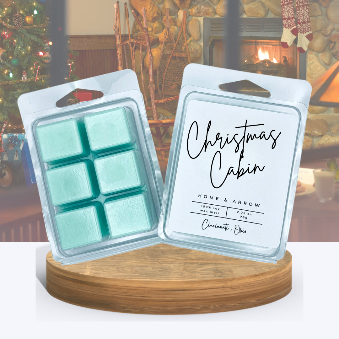 Christmas Cabin green wax melts in clamshell packaging, displayed on a wooden stand over a cabin room scene with a crackling fire and presents under the Christmas tree.