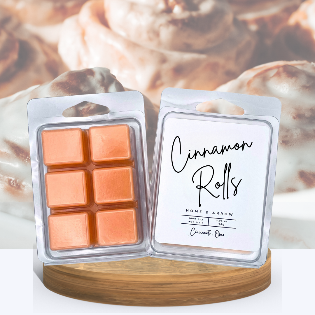 Cinnamon Rolls soy wax melts in clamshell packaging, displayed on a wooden stand with warm cinnamon rolls in the background.