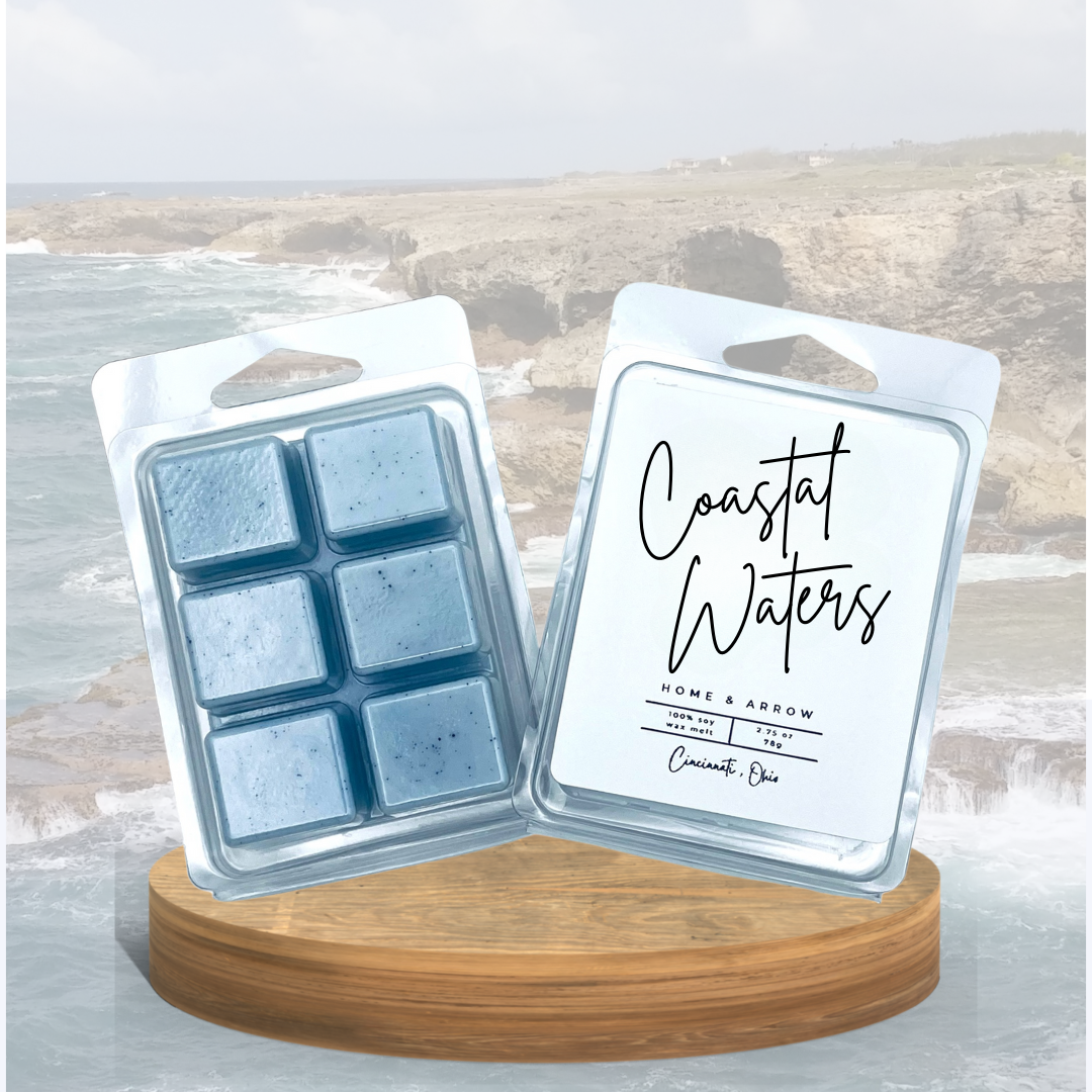 Coastal Waters blue wax melts in clamshell packaging, displayed on a wooden stand showing a rocky coastline in the background.