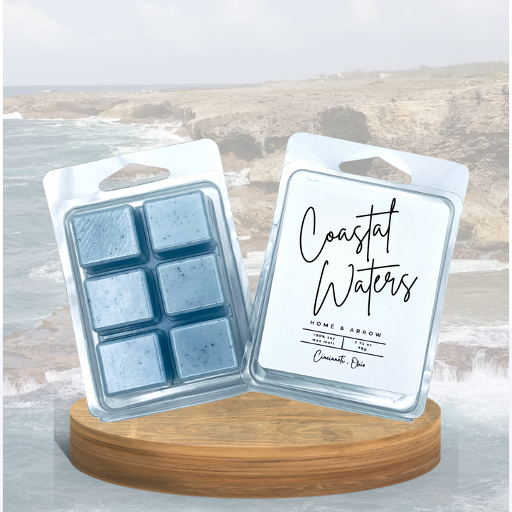 Coastal Waters blue wax melts in clamshell packaging, displayed on a wooden stand showing a rocky coastline in the background.