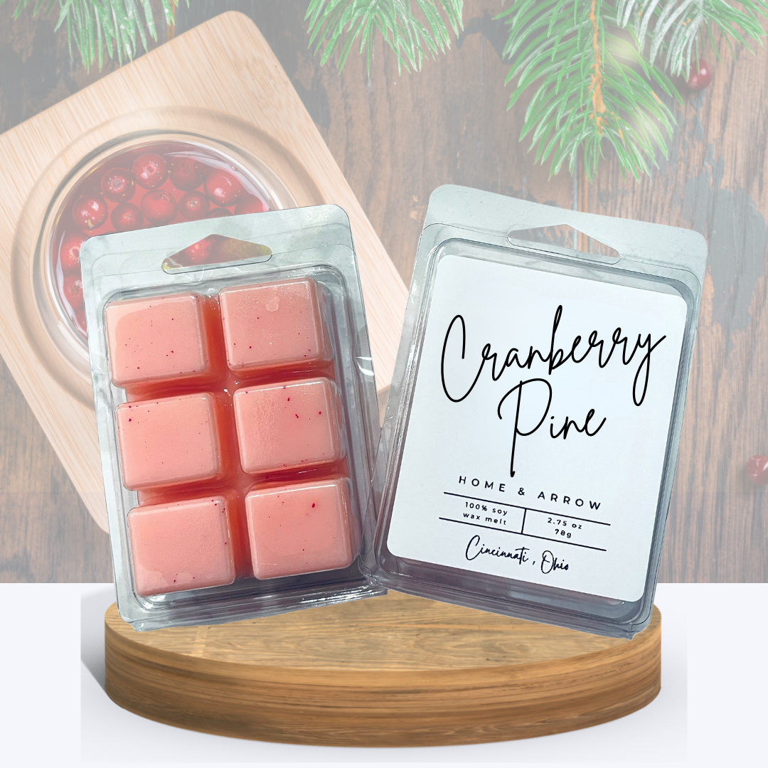 Cranberry Pine soy wax melts in clamshell packaging, displayed on a wooden stand a bowl of cranberries on a serving board with pine surrounding it.