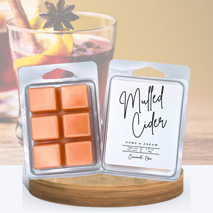 Mulled Cider orange wax melts in clamshell packaging, displayed on a wooden stand with several almond colored macaron cookies in the background.