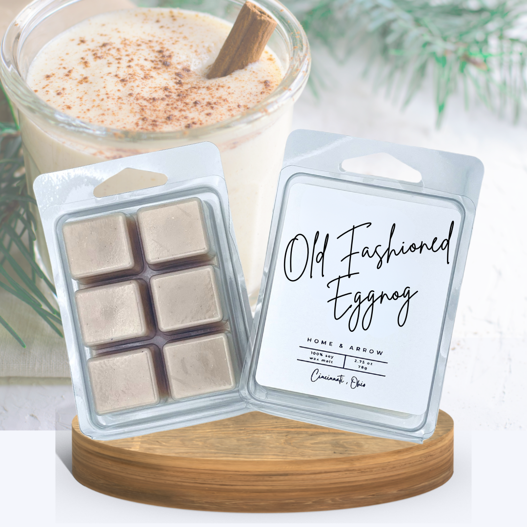 Old Fashioned Eggnog wax melts in clamshell packaging displayed on a wooden stand with a festive glass of eggnog dipped with a cinnamon stick in the background.