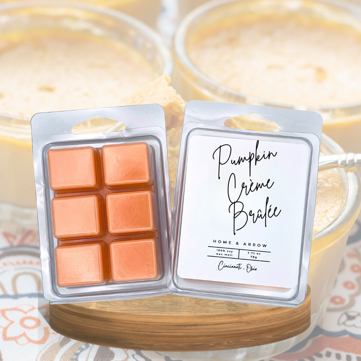 Two wax melts packages displayed on a wooden coaster in front of a softly blurred background of pumpkin creme brulee. The left package contains six orange-brown wax cubes, and the right package is labeled 'Pumpkin Crème Brûlée' by Home & Arrow.