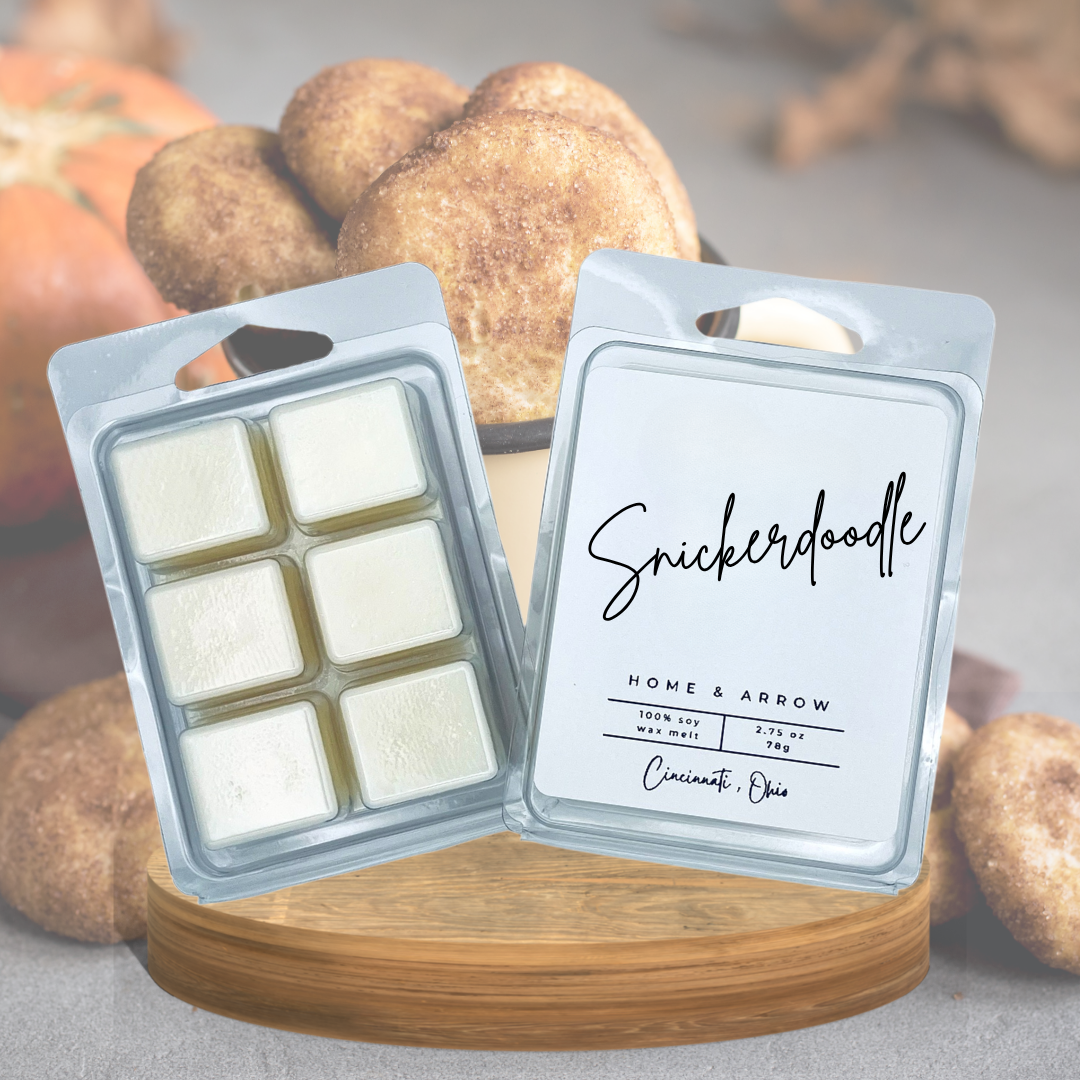 Two Snickerdoodle scented wax melts in clear plastic clamshell packaging, one showing the six white wax cubes inside and the other displaying the front label, set on a wooden block with cinnamon-sugar-coated treats blurred in the background.
