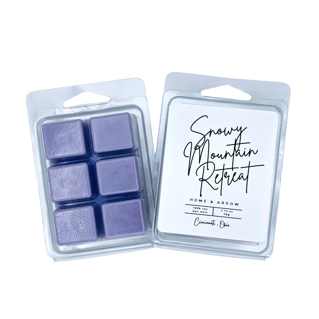 Two clear plastic wax melts packages on a white background. One package shows six lavender-purple wax cubes, and the other shows the white insert card with black script reading 'Snowy Mountain Retreat' and smaller text 'Home & Arrow' and 'Cinnamon Dog'.