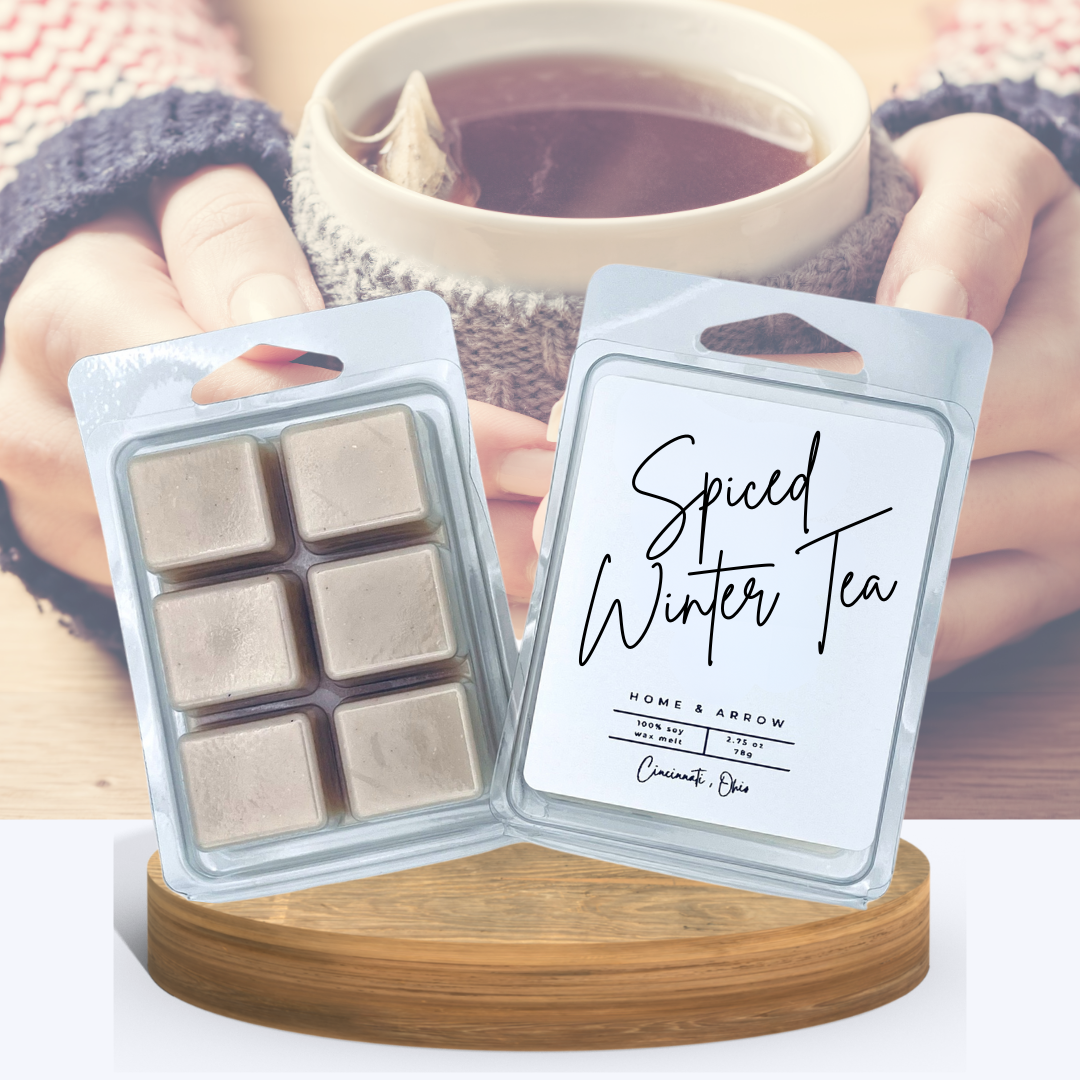 Spiced Winter Tea wax melts in clamshell packaging displayed on a wooden stand with  a woman holding a steaming cup of tea wrapped in a knitted cozy in the background.