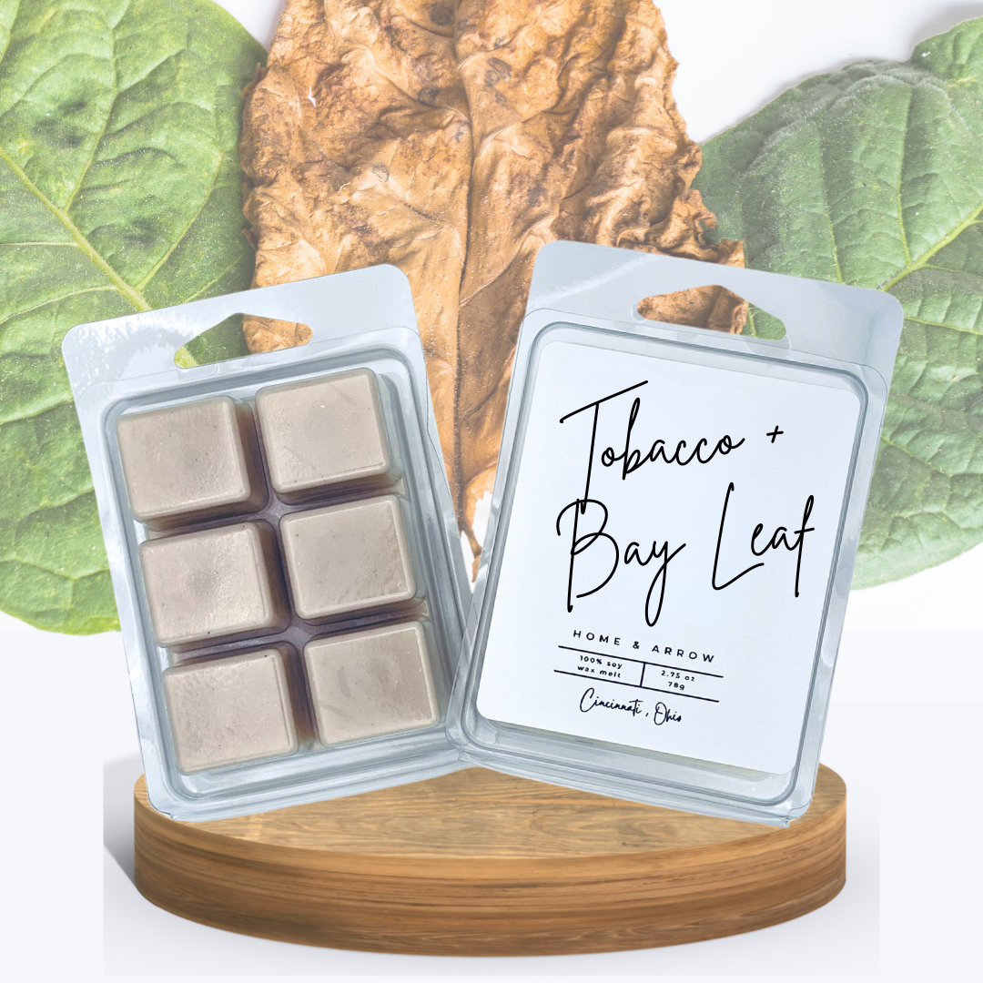 Tobacco + Bay Leaf soy wax melts in clamshell packaging, displayed on a wooden stand with  a brown tobacco leaf and green bay leaves in the background.