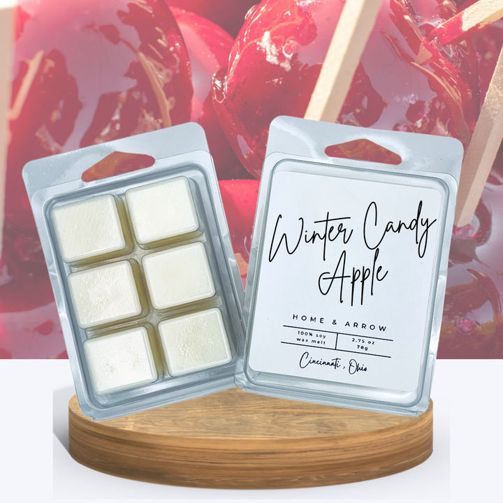 Winter Candy Apple white wax melts in clamshell packaging, displayed on a wooden stand with several almond colored macaron cookies in the background.